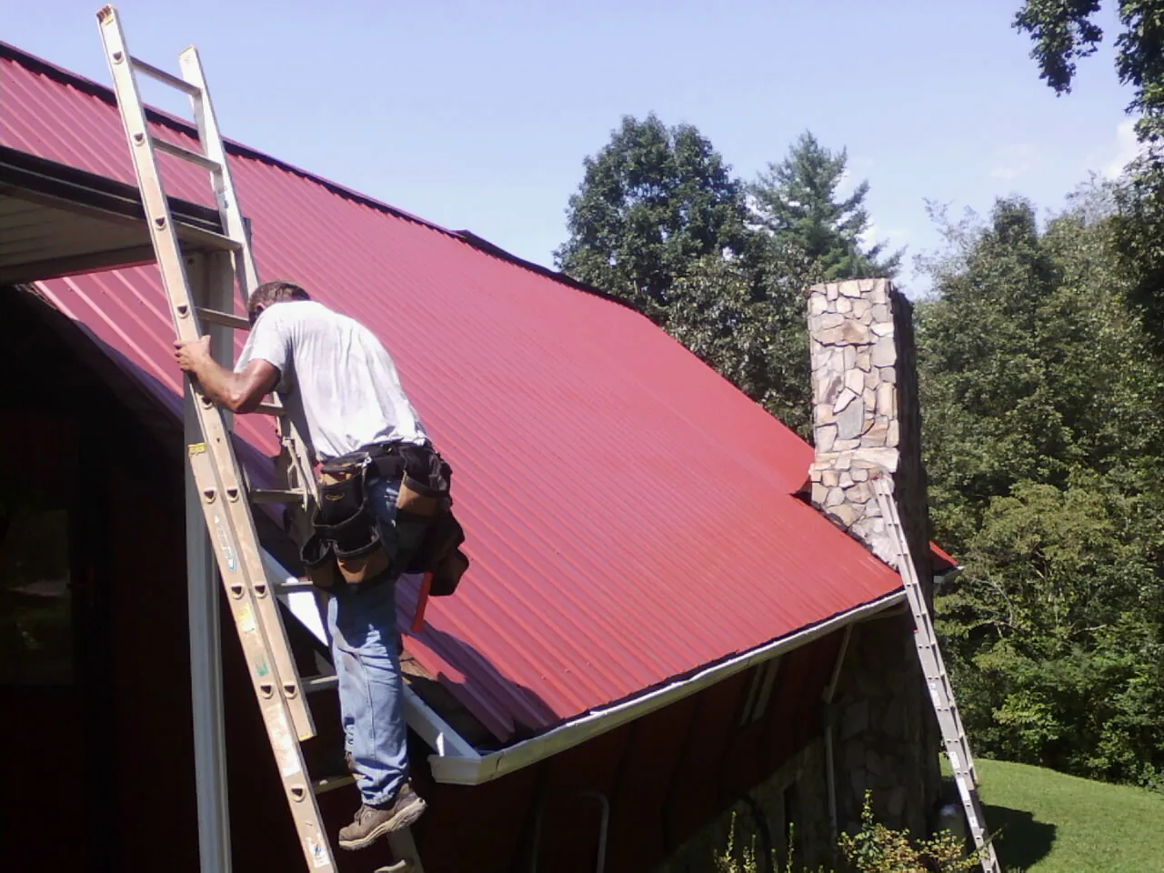 Roofer on a ladder beside a red metal roof and stone chimney — Soffit & Fascia Repair by Roofing Brothers in Locust Gap